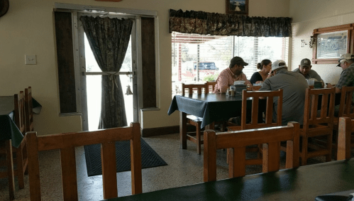 A cozy restaurant interior with wooden tables and chairs, featuring diners at a table and a door leading outside.